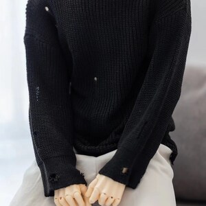 Puede incluir: Un su&eacute;ter de punto negro con detalles desgastados y agujeros por todas partes. El su&eacute;ter tiene mangas largas y un cuello redondo.