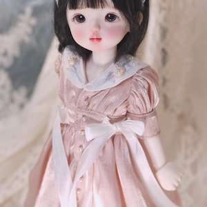 Puede incluir: Una muñeca de porcelana con cabello negro que lleva un vestido rosa con encaje blanco y detalles de perlas. La muñeca lleva una diadema blanca con detalles de perlas y flores.