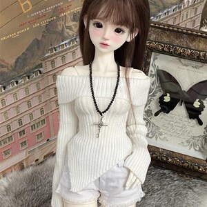 Puede incluir: Una mu&ntilde;eca blanca con cabello largo y casta&ntilde;o que lleva un su&eacute;ter blanco de hombros descubiertos con un collar de cuentas negras y un colgante de cruz plateada. La mu&ntilde;eca tambi&eacute;n lleva pantalones cortos de mezclilla blancos.