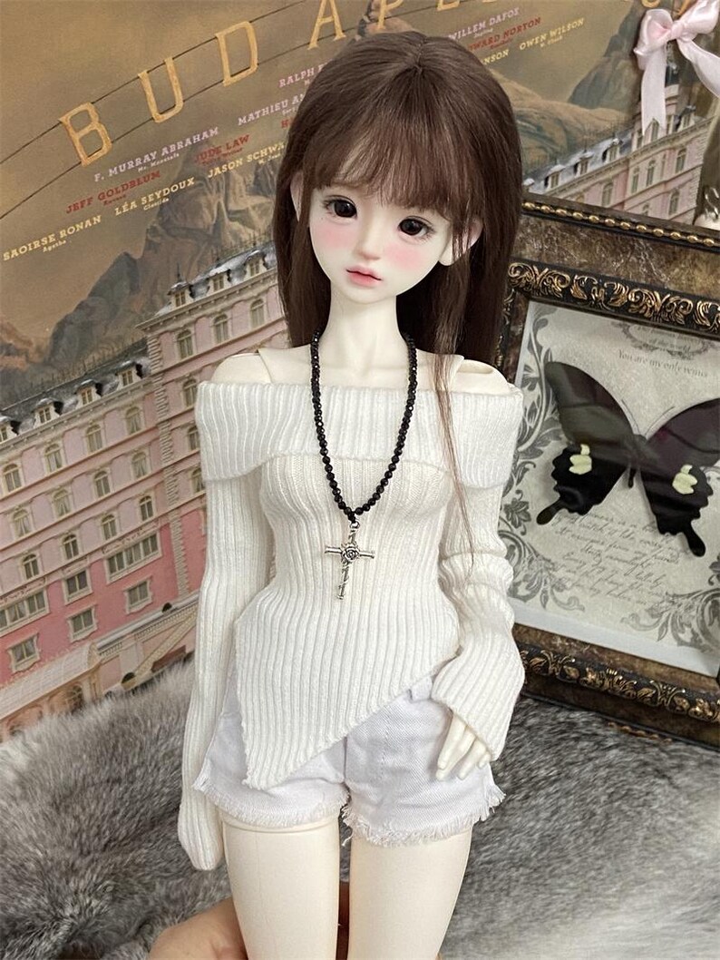 Puede incluir: Una mu&ntilde;eca blanca con cabello largo y casta&ntilde;o que lleva un su&eacute;ter blanco de hombros descubiertos con un collar de cuentas negras y un colgante de cruz plateada. La mu&ntilde;eca tambi&eacute;n lleva pantalones cortos de mezclilla blancos.