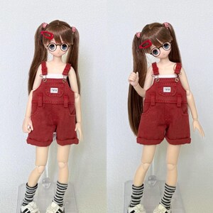 May include: Two dolls wearing red overalls with the text "TWW" on the bib. The dolls have brown hair in pigtails, round glasses, and striped socks with white and gray sneakers. The background is plain white.
