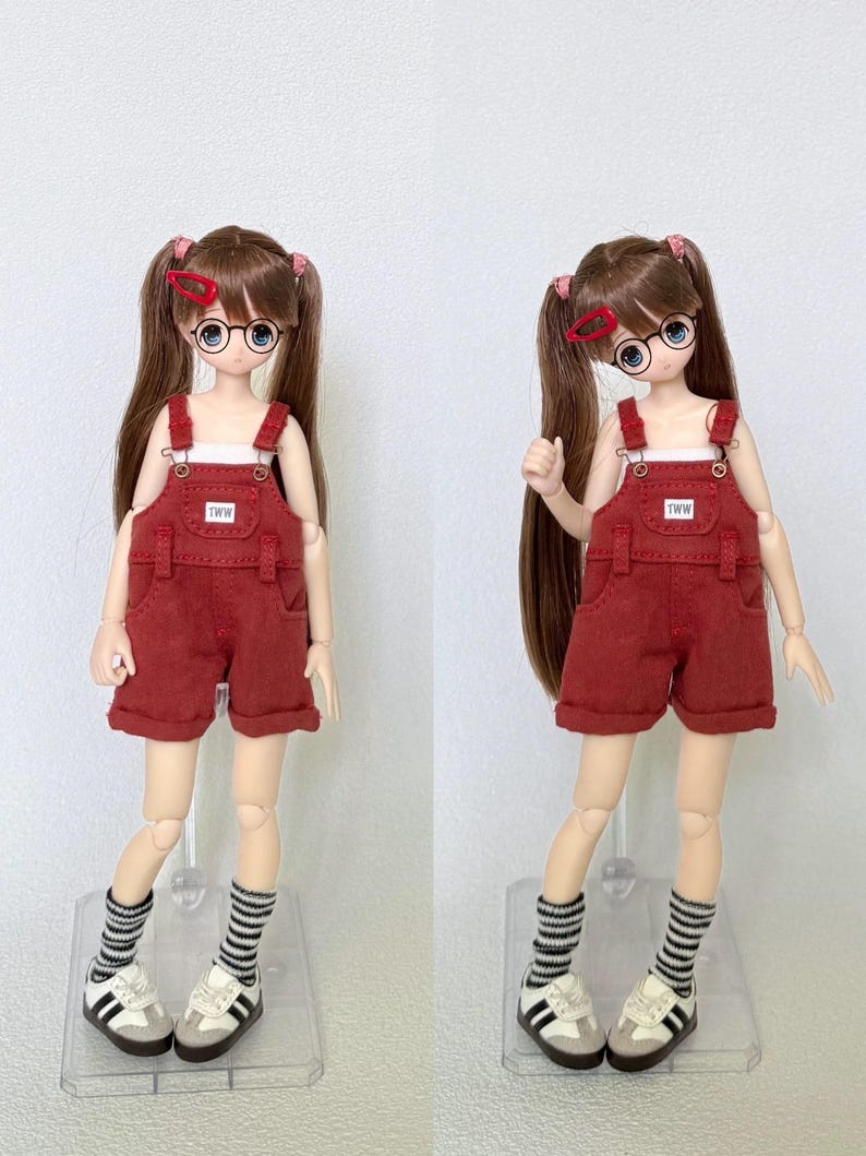 May include: Two dolls wearing red overalls with the text "TWW" on the bib. The dolls have brown hair in pigtails, round glasses, and striped socks with white and gray sneakers. The background is plain white.