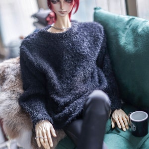 May include: A doll with red hair wearing a dark blue fuzzy sweater and black trousers, seated on a green cushion. The doll has a silver necklace and ring. A black mug with a white design sits nearby.