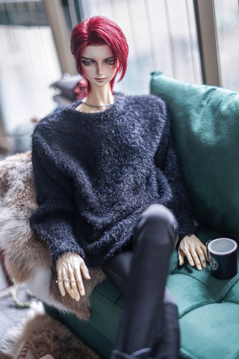 May include: A doll with red hair wearing a dark blue fuzzy sweater and black trousers, seated on a green cushion. The doll has a silver necklace and ring. A black mug with a white design sits nearby.