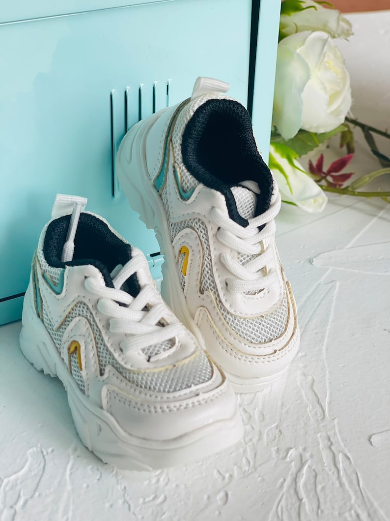 Puede incluir: Un par de zapatillas blancas con detalles negros y un dise&ntilde;o de malla. Las zapatillas tienen una suela gruesa y un acento azul claro en el lateral.