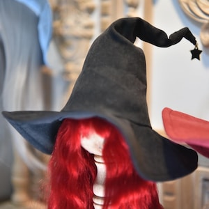 Puede incluir: Un sombrero de bruja negro con ala ancha y punta rizada, adornado con un pequeño dije de estrella. El sombrero se asienta sobre un soporte de madera con pelo largo y rojo vibrante, creando un aspecto caprichoso y festivo.