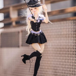 May include: A doll dressed in a black uniform with a matching hat and boots. The doll is holding a riding crop and has long blonde hair. The outfit includes a pleated skirt, vest, and a belt. The doll is posed in a dynamic stance.
