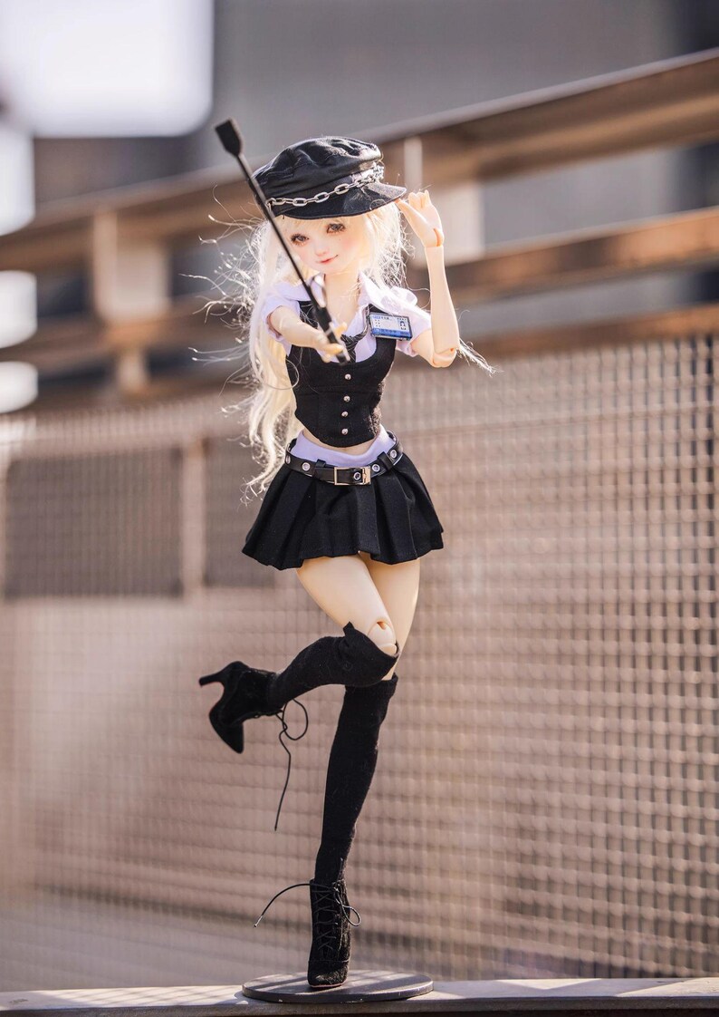 May include: A doll dressed in a black uniform with a matching hat and boots. The doll is holding a riding crop and has long blonde hair. The outfit includes a pleated skirt, vest, and a belt. The doll is posed in a dynamic stance.