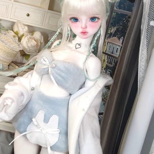 May include: A white doll with long blonde hair styled in braids with blue accents. The doll is wearing a light blue fuzzy top and matching shorts with a white bunny design. The doll is also wearing a white jacket.