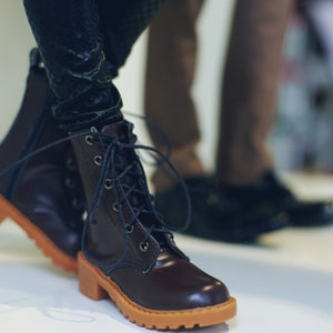 May include: Brown leather boots with orange soles and laces. The boots have a classic design with a round toe and a sturdy heel. The laces are dark blue and the boots are worn with dark pants.