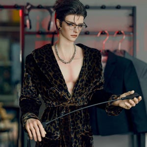 May include: A doll wearing a leopard print robe with a black belt and holding a black riding crop.