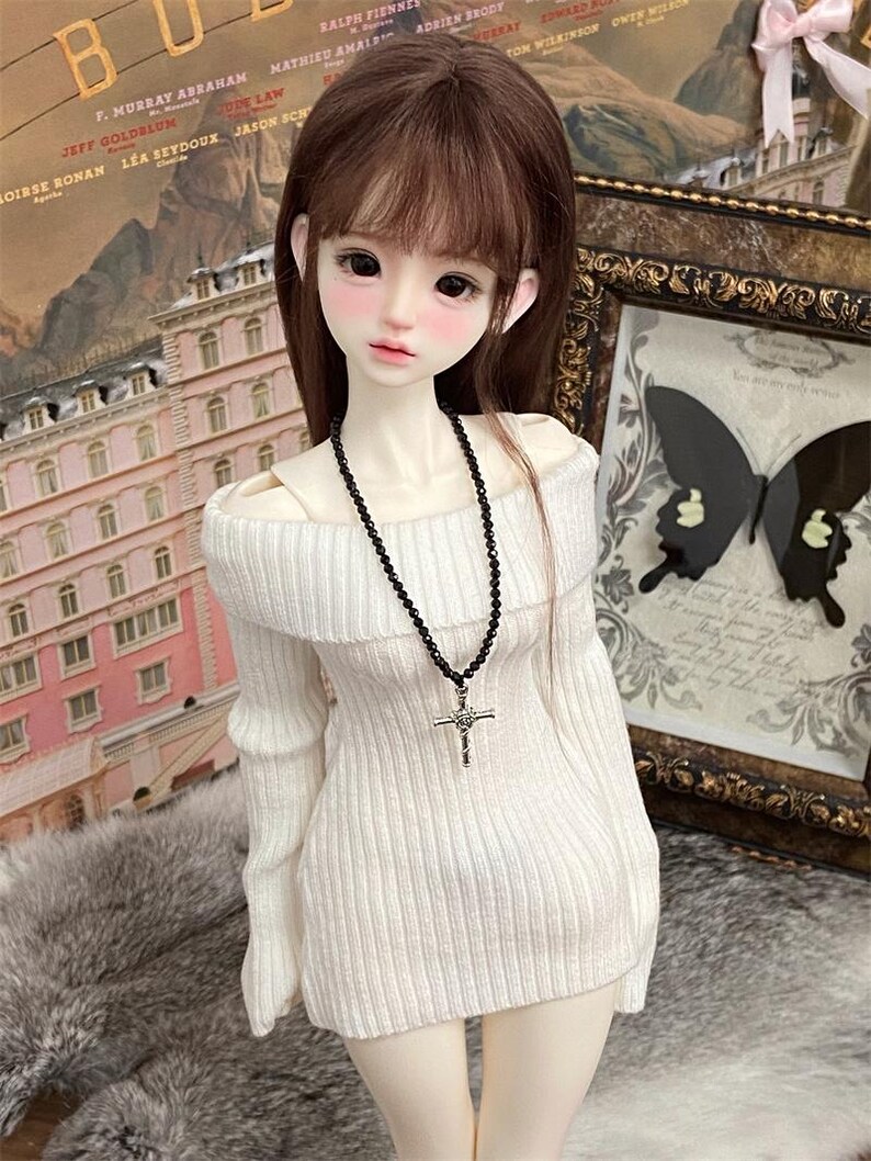 Puede incluir: Un su&eacute;ter blanco de hombros descubiertos con mangas largas y textura acanalada. El su&eacute;ter lo lleva una mu&ntilde;eca con el pelo largo y casta&ntilde;o y un collar de cuentas negras con un colgante de cruz plateada.