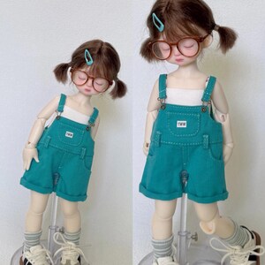 May include: Two dolls wearing teal denim overalls with white tops, glasses, and sneakers. The overalls have the text "TWW" on the pocket. The dolls have brown hair in pigtails and are wearing gray and white striped socks.