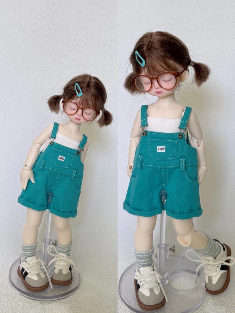 May include: Two dolls wearing teal denim overalls with white tops, glasses, and sneakers. The overalls have the text "TWW" on the pocket. The dolls have brown hair in pigtails and are wearing gray and white striped socks.