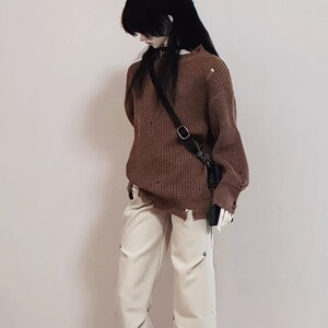 Puede incluir: Un su&eacute;ter de punto marr&oacute;n desgastado con una bolsa de correa negra que se lleva sobre el hombro. El su&eacute;ter se combina con pantalones blancos.