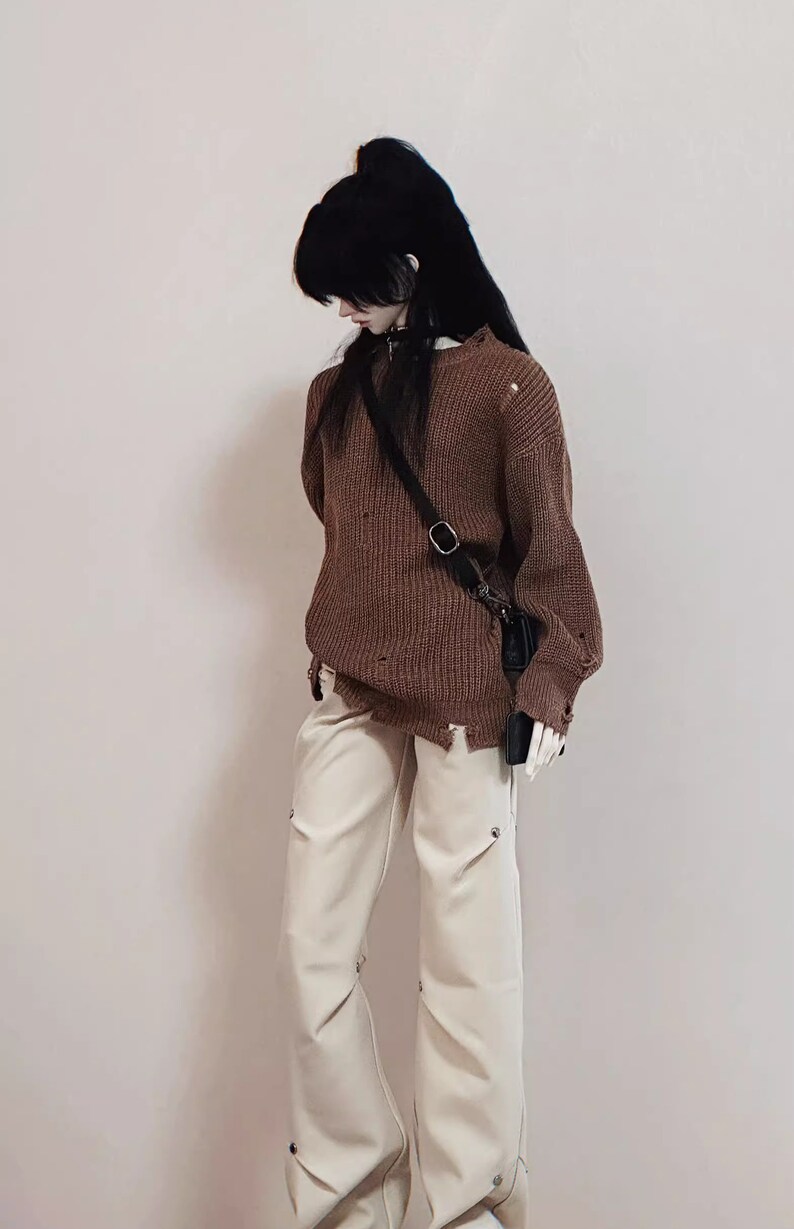 Puede incluir: Un su&eacute;ter de punto marr&oacute;n desgastado con una bolsa de correa negra que se lleva sobre el hombro. El su&eacute;ter se combina con pantalones blancos.