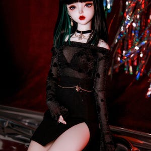 May include: A black and green doll with long black hair and a black outfit with a sheer top and a skirt with a slit. The doll is wearing a black choker necklace with a silver pendant and black stockings.