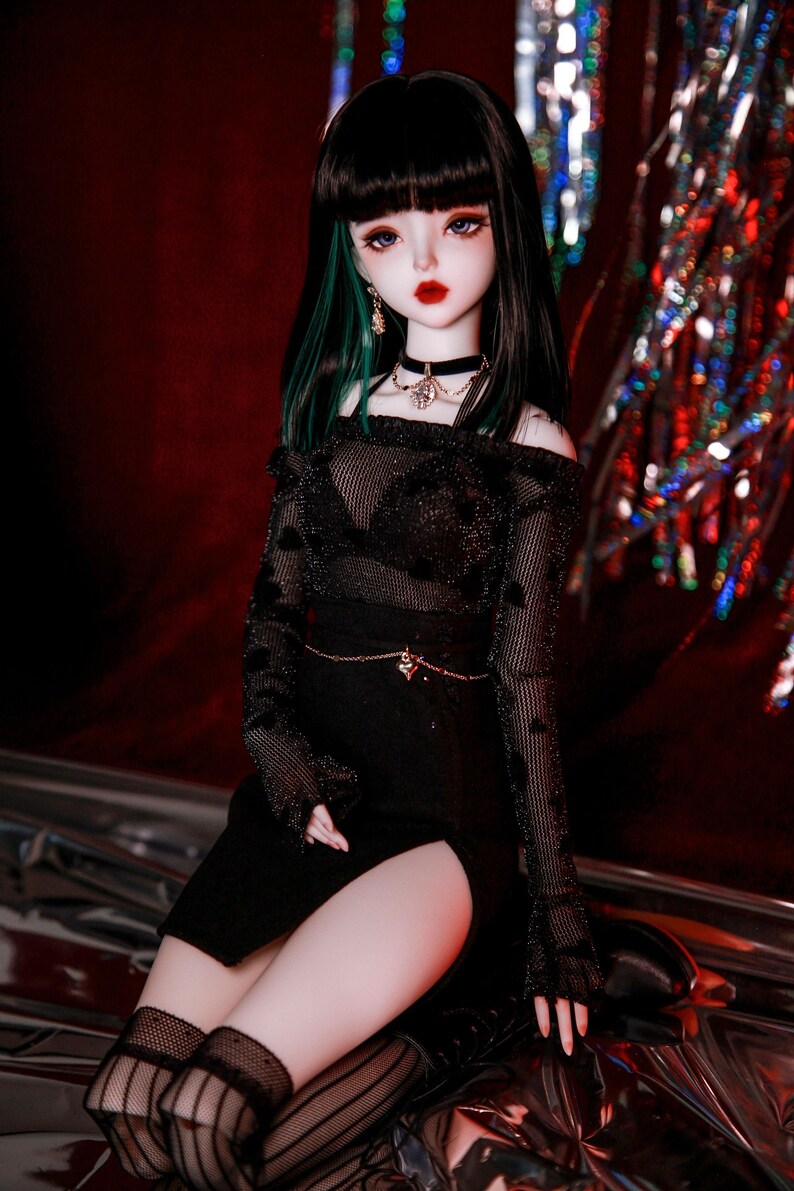 May include: A black and green doll with long black hair and a black outfit with a sheer top and a skirt with a slit. The doll is wearing a black choker necklace with a silver pendant and black stockings.