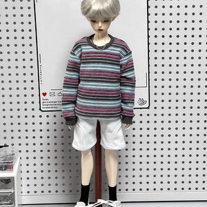 Pu&ograve; includere: Una bambola che indossa una camicia a maniche lunghe a righe con colletto bianco, pantaloncini bianchi e scarpe da ginnastica bianche. La bambola ha i capelli corti e argentati ed &egrave; in piedi su un supporto di legno. Lo sfondo &egrave; un pannello forato bianco.