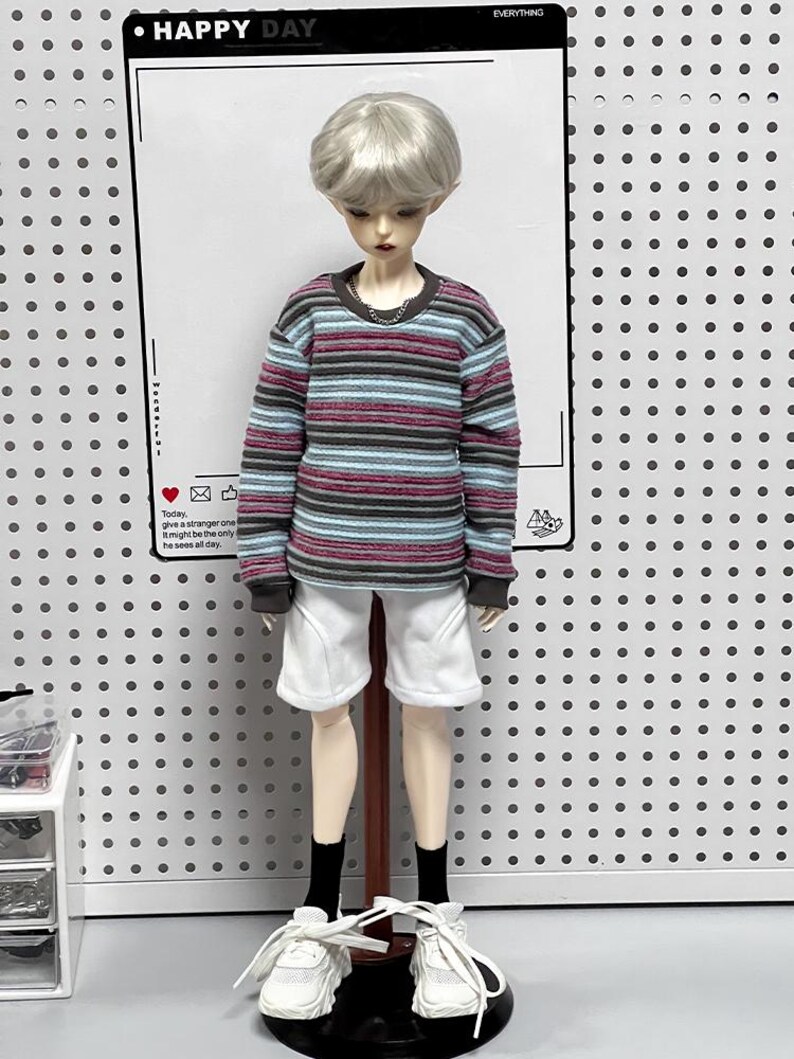 Pu&ograve; includere: Una bambola che indossa una camicia a maniche lunghe a righe con colletto bianco, pantaloncini bianchi e scarpe da ginnastica bianche. La bambola ha i capelli corti e argentati ed &egrave; in piedi su un supporto di legno. Lo sfondo &egrave; un pannello forato bianco.