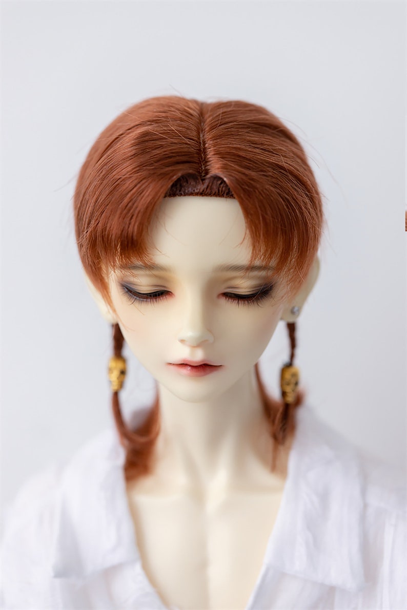 May include: A doll with long, auburn hair styled in two braids with skull-shaped charms. The doll is wearing a white collared shirt.
