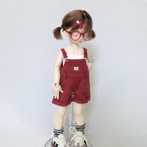 May include: A doll wearing a red overall shorts set with a white top, brown glasses, and sneakers. The shorts have a small pocket with the letters "TWW". The doll has brown hair in pigtails and is wearing striped socks.