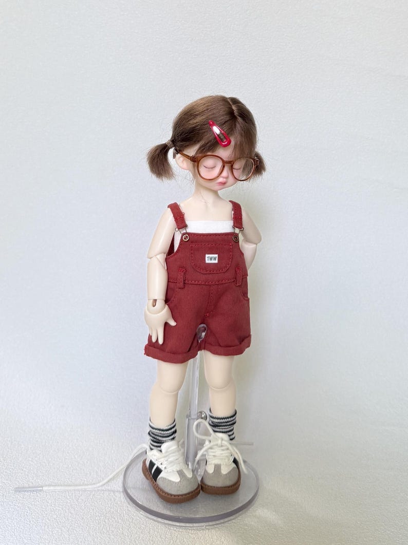 May include: A doll wearing a red overall shorts set with a white top, brown glasses, and sneakers. The shorts have a small pocket with the letters "TWW". The doll has brown hair in pigtails and is wearing striped socks.