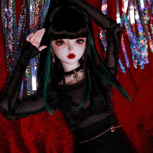 May include: A doll with long black hair and green highlights wearing a black mesh top with a black skirt and a silver chain belt. The doll has blue eyes and red lipstick. The doll is posed against a red background with silver and red streamers.