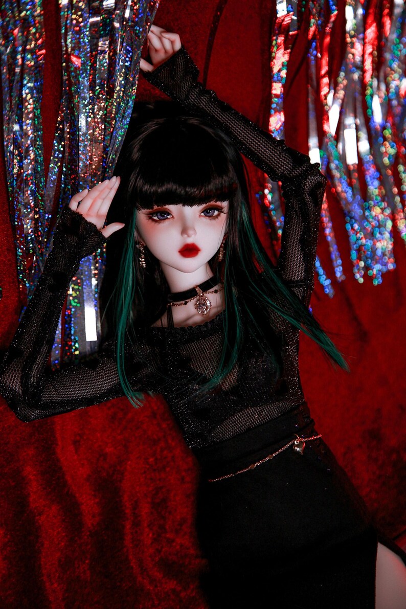 May include: A doll with long black hair and green highlights wearing a black mesh top with a black skirt and a silver chain belt. The doll has blue eyes and red lipstick. The doll is posed against a red background with silver and red streamers.