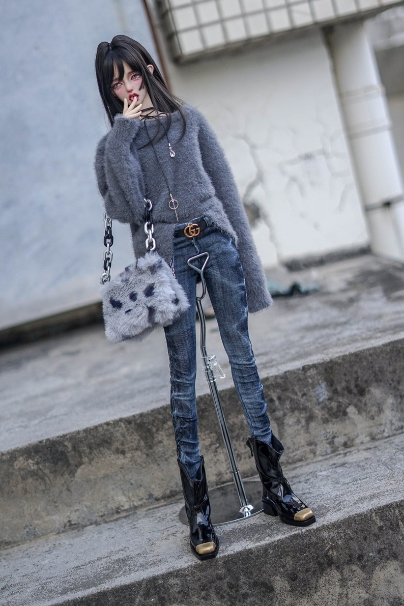 May include: A doll in a grey fluffy jumper, blue jeans, and black boots with gold details. The doll carries a small grey bag with a chain strap and a black belt with a gold buckle. The doll is standing on a concrete surface.