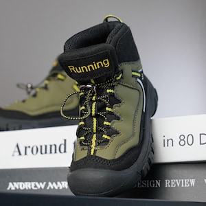 Può includere: Uno scarponcino da trekking verde oliva e nero con dettagli gialli. La parola "Running" è stampata in giallo sulla linguetta. Lo scarponcino ha lacci neri con dettagli gialli e una suola nera. Un altro scarponcino è visibile sullo sfondo.
