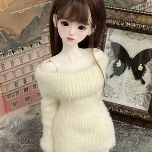 Puede incluir: Una mu&ntilde;eca con cabello largo y casta&ntilde;o que lleva un vestido de su&eacute;ter blanco y esponjoso con hombros descubiertos. La mu&ntilde;eca tiene una tez clara y lleva un maquillaje ligero. La mu&ntilde;eca est&aacute; de pie frente a una pared con un p&oacute;ster y una imagen enmarcada.