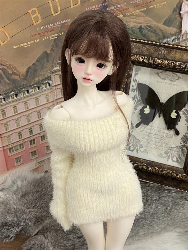 Puede incluir: Una mu&ntilde;eca con cabello largo y casta&ntilde;o que lleva un vestido de su&eacute;ter blanco y esponjoso con hombros descubiertos. La mu&ntilde;eca tiene una tez clara y lleva un maquillaje ligero. La mu&ntilde;eca est&aacute; de pie frente a una pared con un p&oacute;ster y una imagen enmarcada.