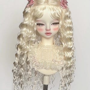 May include: A ball-jointed doll head featuring long, wavy, platinum blonde hair styled with two small pink bows on each side. The doll has a pale complexion with rosy pink cheeks, dark eyelashes, and wears a cream-coloured lace collar with ruffles.