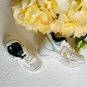 Puede incluir: Un par de zapatillas blancas con detalles en azul y amarillo. Las zapatillas tienen una parte superior de malla y una suela gruesa.