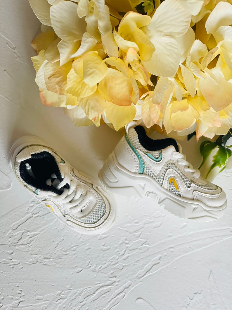 Puede incluir: Un par de zapatillas blancas con detalles en azul y amarillo. Las zapatillas tienen una parte superior de malla y una suela gruesa.
