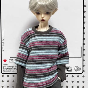 Pu&ograve; includere: Una bambola che indossa un maglione a righe con maniche lunghe e pantaloni grigi. Il maglione ha righe rosa, blu e grigie. La bambola ha i capelli corti e argentati e guarda in basso.