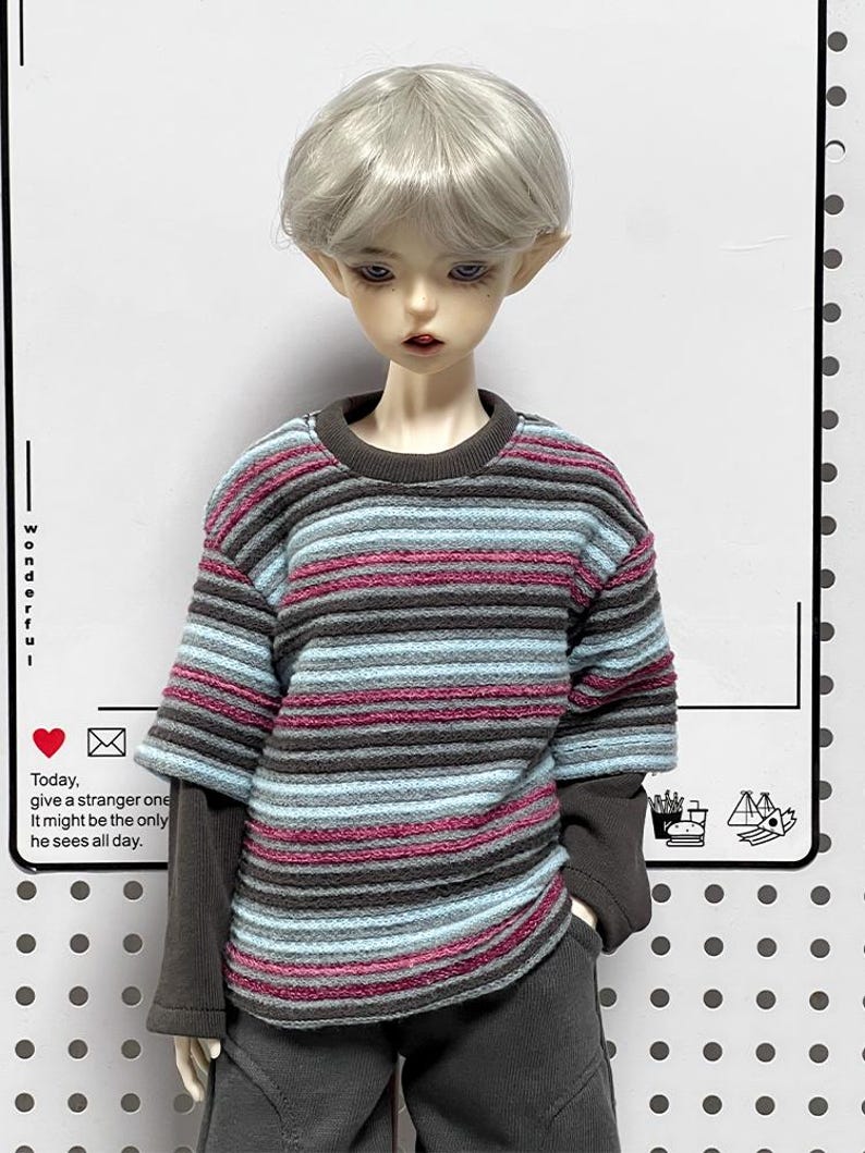 Pu&ograve; includere: Una bambola che indossa un maglione a righe con maniche lunghe e pantaloni grigi. Il maglione ha righe rosa, blu e grigie. La bambola ha i capelli corti e argentati e guarda in basso.