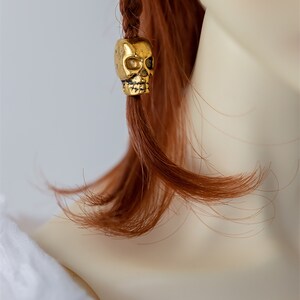May include: A gold skull bead charm for hair. The bead is attached to a braid of red hair.