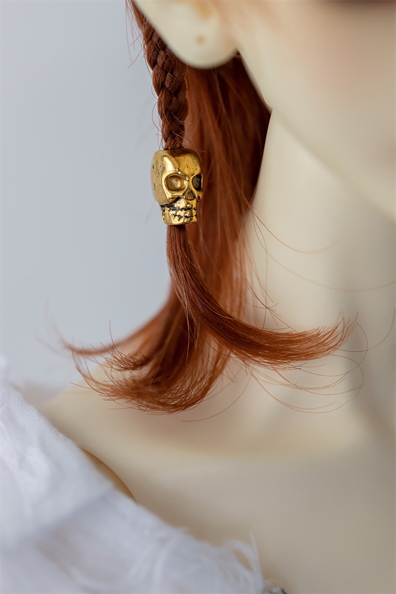 May include: A gold skull bead charm for hair. The bead is attached to a braid of red hair.