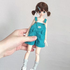 May include: A doll with brown hair styled in pigtails, wearing a teal overall shorts set. The doll has blue eyes and is wearing white sneakers with black stripes and gray and white striped socks. The overall shorts have the letters "TWW" on the front.