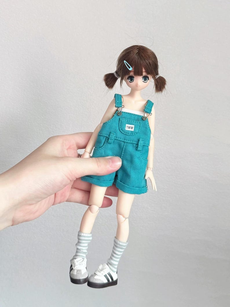 May include: A doll with brown hair styled in pigtails, wearing a teal overall shorts set. The doll has blue eyes and is wearing white sneakers with black stripes and gray and white striped socks. The overall shorts have the letters "TWW" on the front.