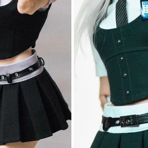May include: A doll wearing a school uniform consisting of a black pleated skirt, a black corset-style top, and a white collared shirt. The outfit includes a black belt and a name tag. The uniform is designed with a focus on detail and style.