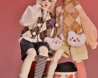 May include: Two dolls wearing winter outfits and hats are sitting on a table with two cups of coffee. The dolls are wearing plaid vests, white sweaters, and white boots. The dolls are holding small plush bags. The cups of coffee have peach designs.