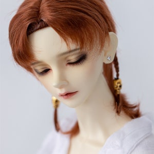 May include: A doll with long, auburn hair styled in two braids with gold accents. The doll is wearing a white shirt and has a neutral expression.