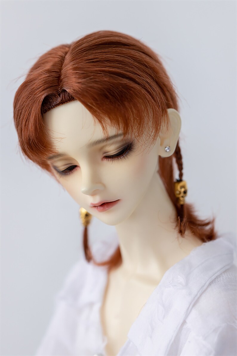 May include: A doll with long, auburn hair styled in two braids with gold accents. The doll is wearing a white shirt and has a neutral expression.