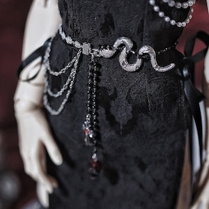 Op de afbeelding: Een zwarte jurk met een zilveren slangenriem, zilveren kettingen en een zwarte kralenketting met rode accenten.