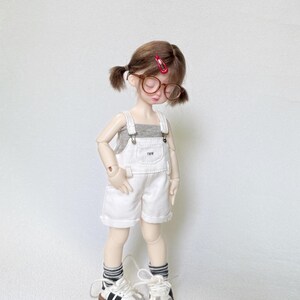 May include: A doll wearing a gray tank top, white overalls, and sneakers. The doll has brown hair styled in pigtails, round glasses, and a red hair clip. The overalls have the letters "TWW" on the front.