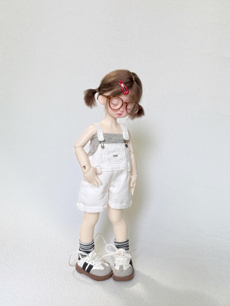May include: A doll wearing a gray tank top, white overalls, and sneakers. The doll has brown hair styled in pigtails, round glasses, and a red hair clip. The overalls have the letters "TWW" on the front.