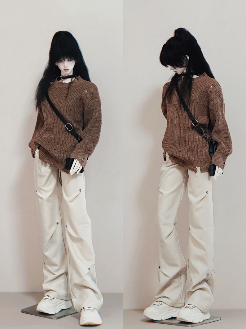 Puede incluir: Una mu&ntilde;eca con un su&eacute;ter marr&oacute;n desgastado con una correa negra y una peque&ntilde;a bolsa negra, pantalones blancos con detalles plateados y zapatillas blancas.